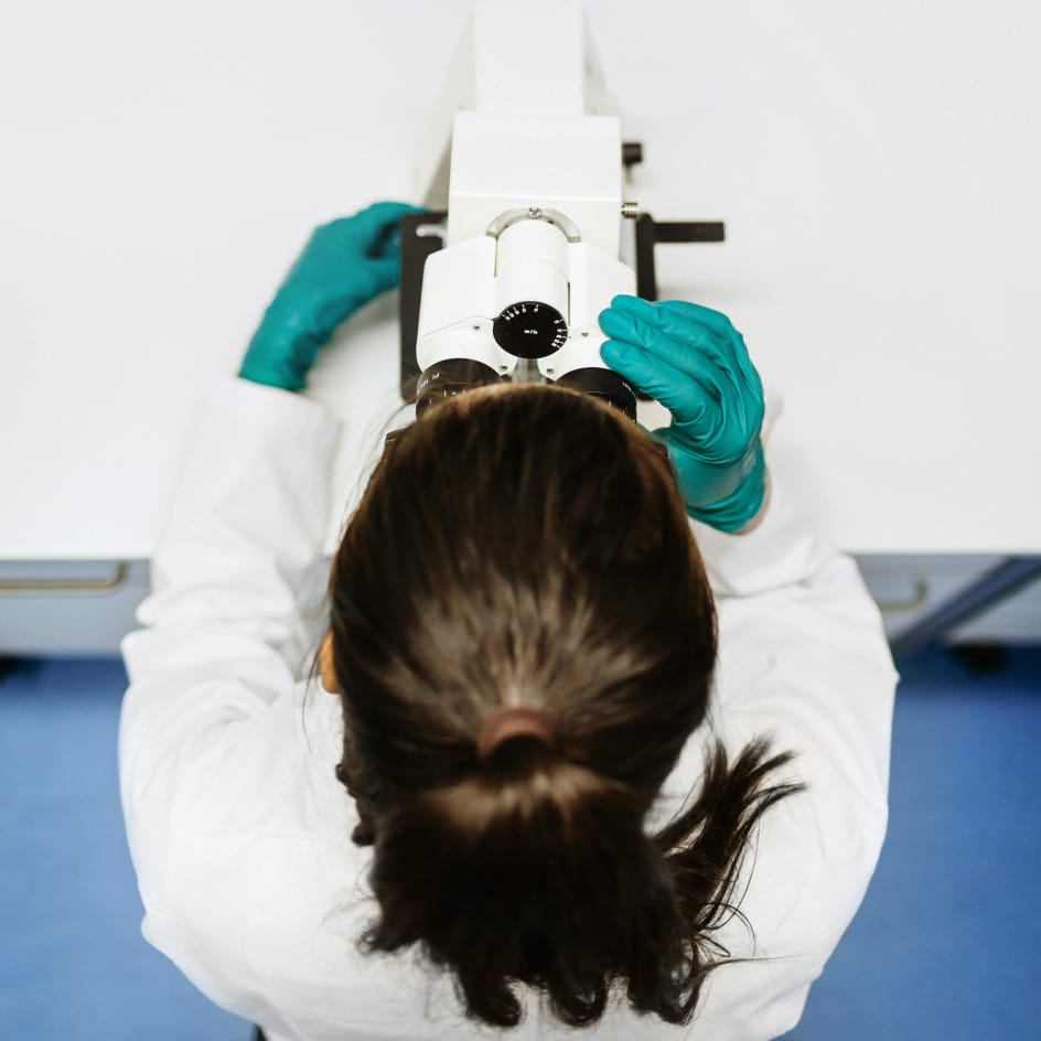 Birdeye view of femal scientist wearing rubber gloves and looking through a white microscope