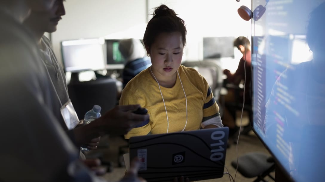 Woman working on a laptop in office with peers and large screen monitor surrounding her 