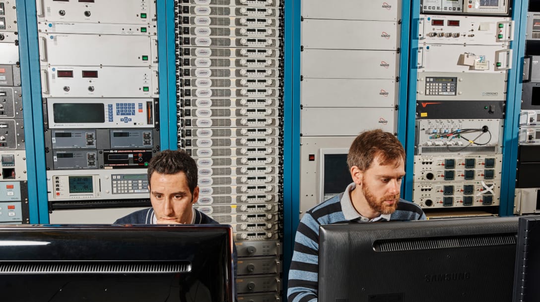 Two scientists working in front of computer monitors with wall of computing equipment behind them