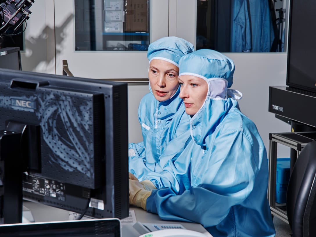 Two female lab workers in blue coveralls and rubber gloves looking at a computer monitor